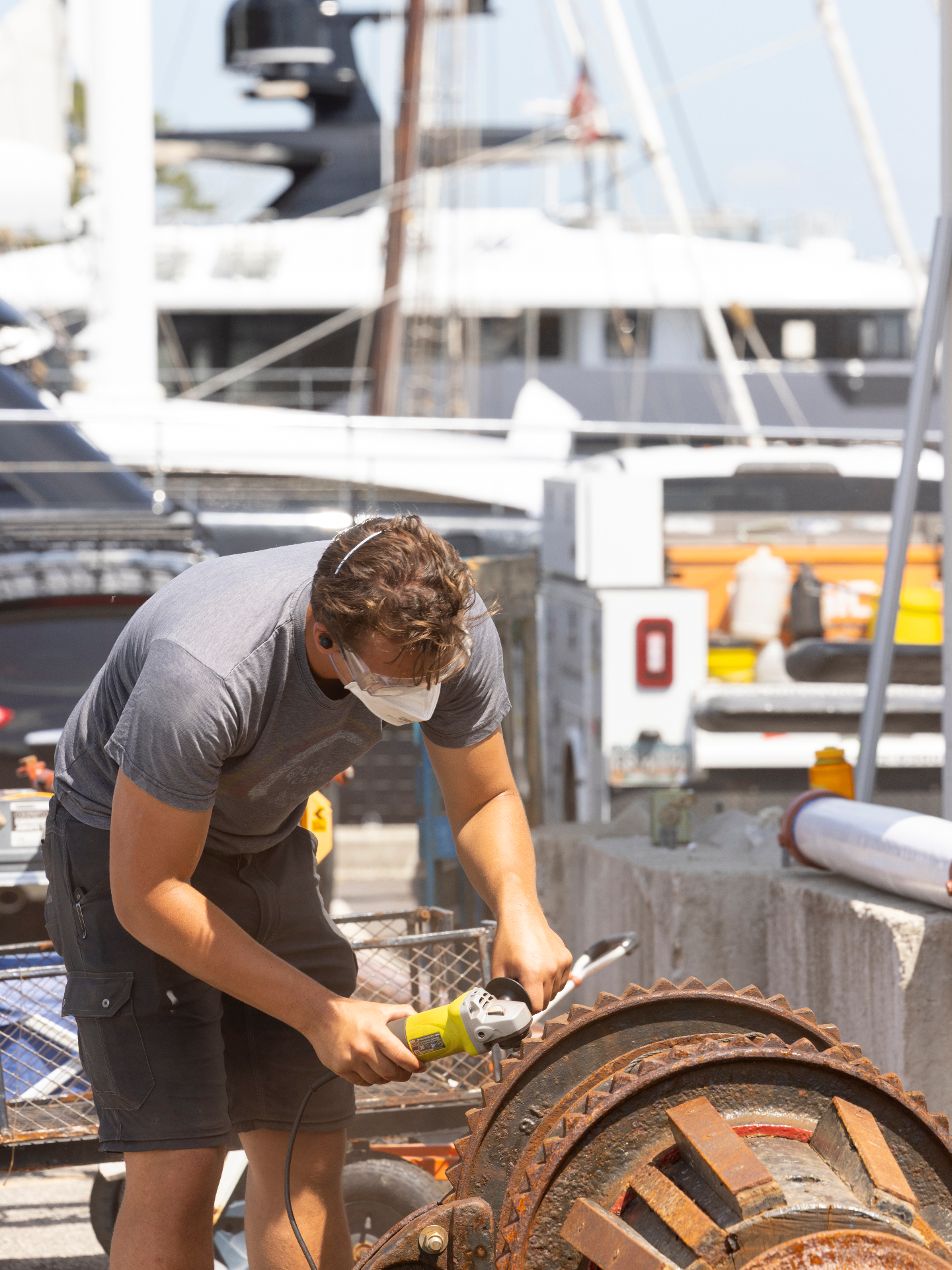 Technician using a grinder on heavy marine equipment near docked yachts.